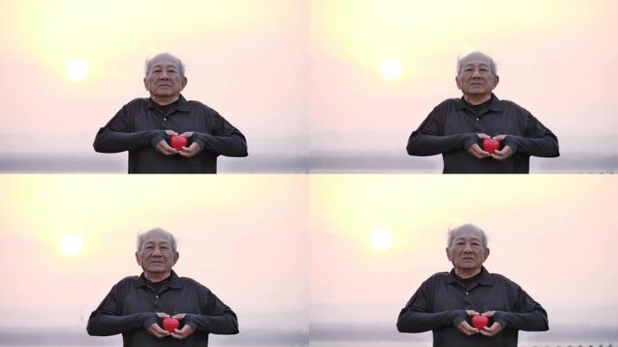 男性前辈抱着红色的心，早晨带着幸福的微笑，日出在后面。健康保健，健康运动和健康的良好开端，保险，世界