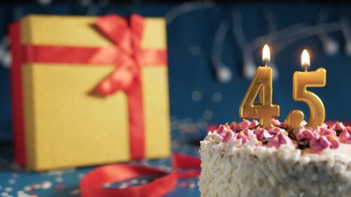 白色生日蛋糕编号45点灯燃烧的金色蜡烛，带灯的蓝色背景和用红丝带绑起来的礼物黄色盒子。特写