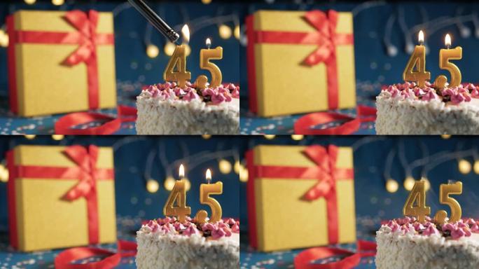 白色生日蛋糕编号45点灯燃烧的金色蜡烛，带灯的蓝色背景和用红丝带绑起来的礼物黄色盒子。特写