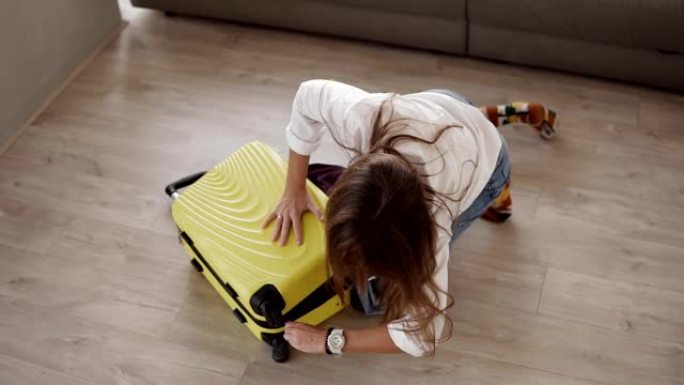 为旅行准备包。年轻女子收拾行李，拼命试图关闭装满的手提箱，坐在上面，关上客厅地板上的拉链。高角度视图