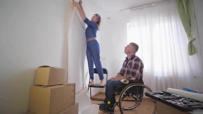 坐在轮椅上的乐于助人的残疾人悬挂照片，而他的妻子用手指做框架，并选择在白色墙壁上拍照的地方