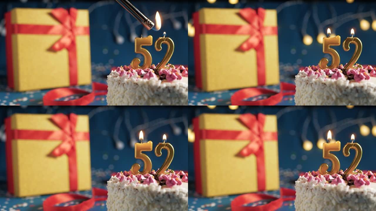 白色生日蛋糕编号52点灯燃烧的金色蜡烛，带灯的蓝色背景和用红丝带绑起来的礼物黄色盒子。特写