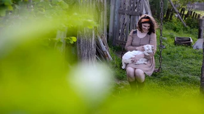 年轻的乡村妇女抱着一只白山羊在乡村院子里的秋千上摇摆。漂亮的大码模特摆姿势。父母之家