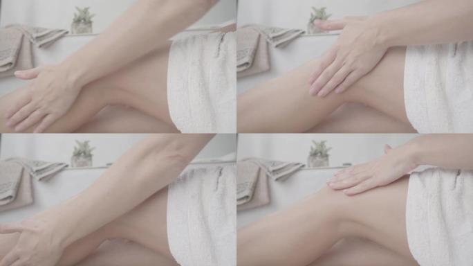 4k平的未分级特写镜头，一个女人在她的设计浴室里，用白毛巾用柔和的心情按摩她的腿