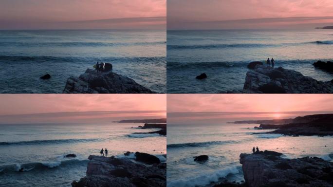 两名女冲浪者站在悬崖/岩石边缘，在日落时观看海浪的美丽航拍。