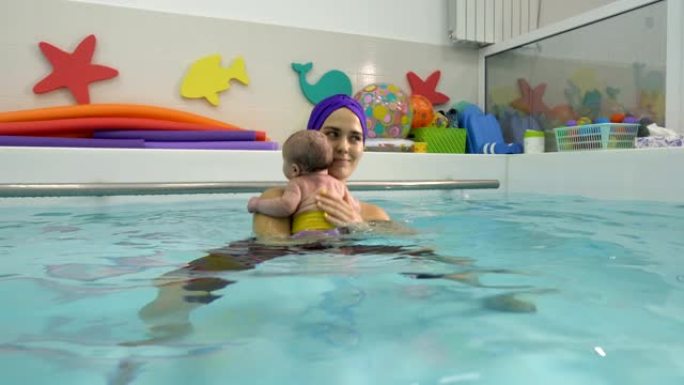 一位快乐的年轻母亲站在儿童游泳池的水中抱着，拥抱和抚摸新生婴儿。婴儿健康在儿童游泳池游泳。母爱。特写