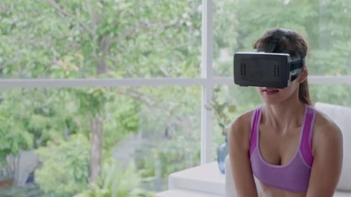 Fit woman用虚拟现实护目镜举重物、壶铃、四处张望、惊喜、兴奋、站立。坐在客厅地板上的亚洲女人
