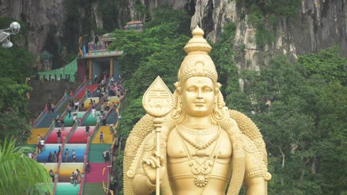 (选择性聚焦) 前景中的Murugan勋爵雕像的壮丽景色，游客爬上彩色楼梯，通往背景中的黑风洞。马来