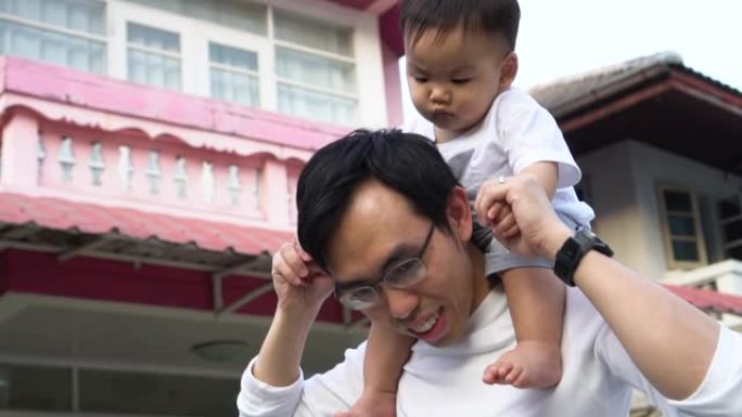 亚洲父亲在房子前面给小儿子背驮。爸爸和孩子享受温暖的家庭时光