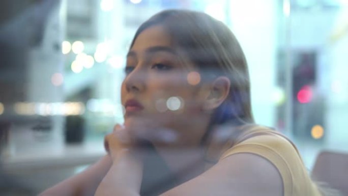 4K机架焦点年轻美丽沮丧的亚洲少女坐在窗前的咖啡馆晚上。孤独悲伤的女人望着窗外的咖啡街，用忧伤的眼神