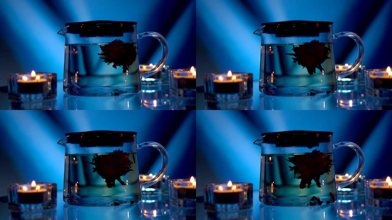 泡茶。绿色的中国茶花在玻璃茶壶中，在蓝色背景下昏暗的灯光下，框架中有蜡烛。放松和安宁的概念