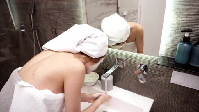一名妇女站在浴室靠近水槽的白色毛巾上，用水龙头上的水洗掉了白色的化妆品面膜。罕见的观点
