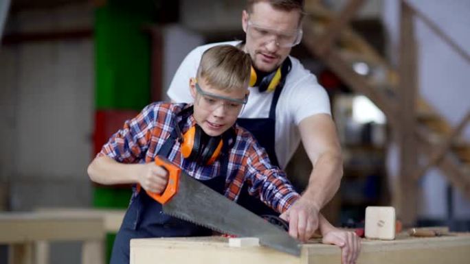 在父亲，专业木匠的帮助下，勤奋的小男孩用手锯切割木板的选择性聚焦镜头，站在木工车间的工作台上