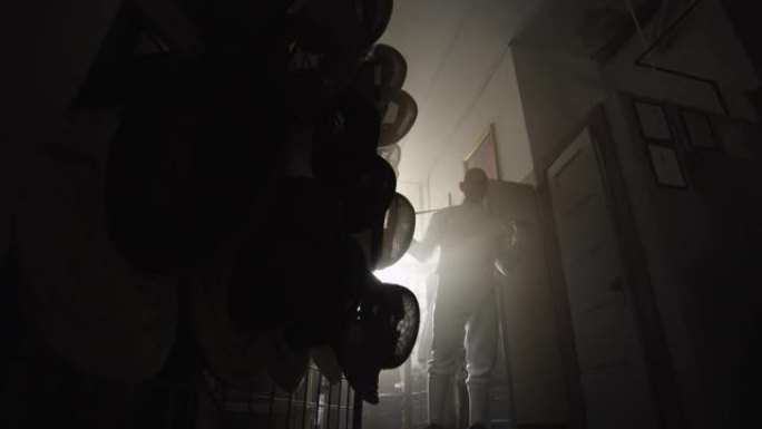 一名男性击剑手在黑暗而尘土飞扬的设备室内用箔纸伸展
