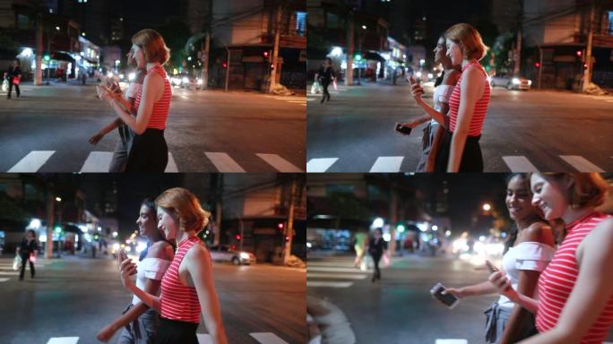 两个女孩多种族的朋友走在夜晚的街道上，展示手机内容屏幕休闲