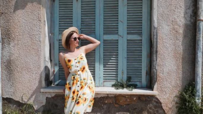 迷人的黑发女人在夏天的连衣裙和太阳眼镜站在附近的老房子与蓝色木制窗户和摆姿势的相机