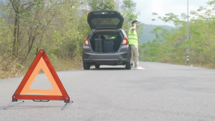 背景乡村道路上的汽车故障后，与担心的亚洲男子通过手机与保险或汽车服务中心公司交谈的道路上的故障三角标
