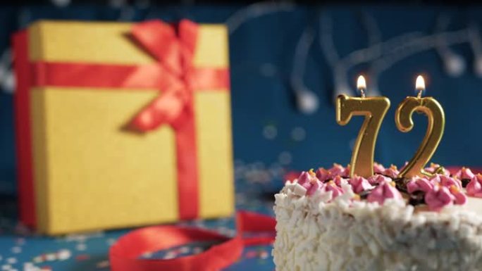 白色生日蛋糕编号72点灯燃烧的金色蜡烛，带灯的蓝色背景和用红丝带绑起来的礼物黄色盒子。特写