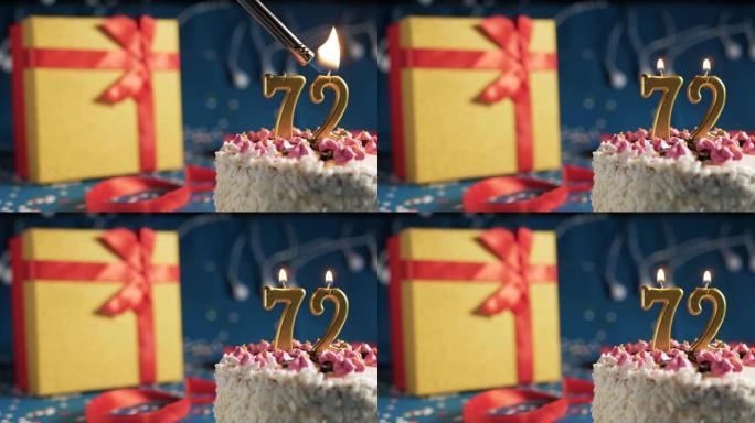 白色生日蛋糕编号72点灯燃烧的金色蜡烛，带灯的蓝色背景和用红丝带绑起来的礼物黄色盒子。特写