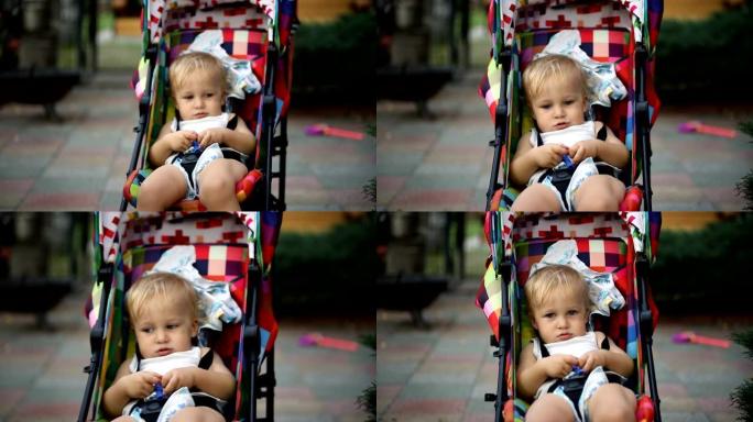 可爱的可爱的高加索金发蹒跚学步的男孩坐在明亮的彩色婴儿车上，在后院散步时被母亲摇晃的肖像
