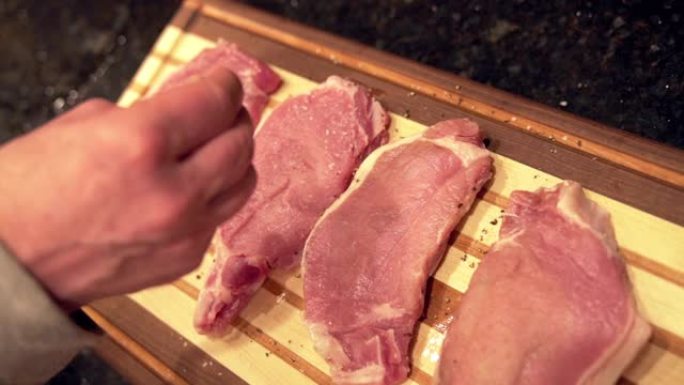 慢动作将粗胡椒粉撒在未煮熟的生去骨里脊肉猪排上，新鲜放在准备烧烤的木制砧板上