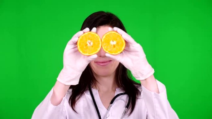年轻女医生的肖像，营养师身着白色医用服，带着听诊器，手里拿着两半橘子，面带微笑。绿色背景