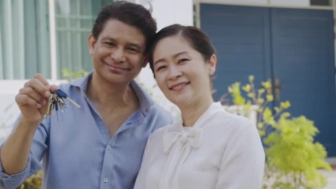 高级夫妇肖像在他们的房子前面拿着房子钥匙。快乐时刻。面对镜头。慢动作。50多岁的亚洲男女。