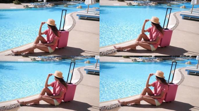 穿着粉红色箱子的年轻女子坐在酒店游泳池附近。旅游度假概念。1920x1080中的高清视频