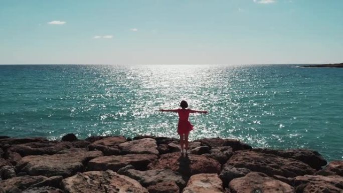 穿着红色连衣裙的女人的剪影，双臂分开在天空中站在珊瑚湾码头的岩石上，看着大海，太阳路在蓝色的水面上倒