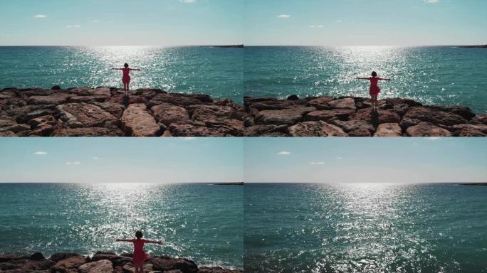 穿着红色连衣裙的女人的剪影，双臂分开在天空中站在珊瑚湾码头的岩石上，看着大海，太阳路在蓝色的水面上倒