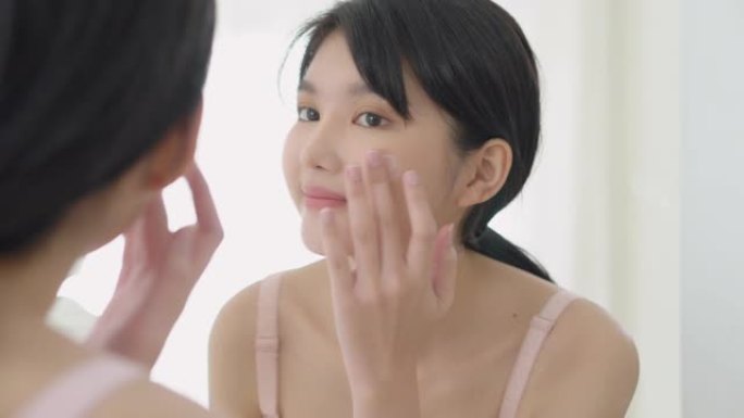 美丽的年轻亚洲女人微笑着看着镜子检查面部和护肤霜和化妆品的年轻化和卫生，美容女孩快乐清洁面部乳液和健