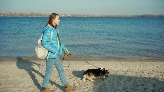 年轻女子和可爱的狗威尔士柯基犬一起沿着海边的沙滩散步