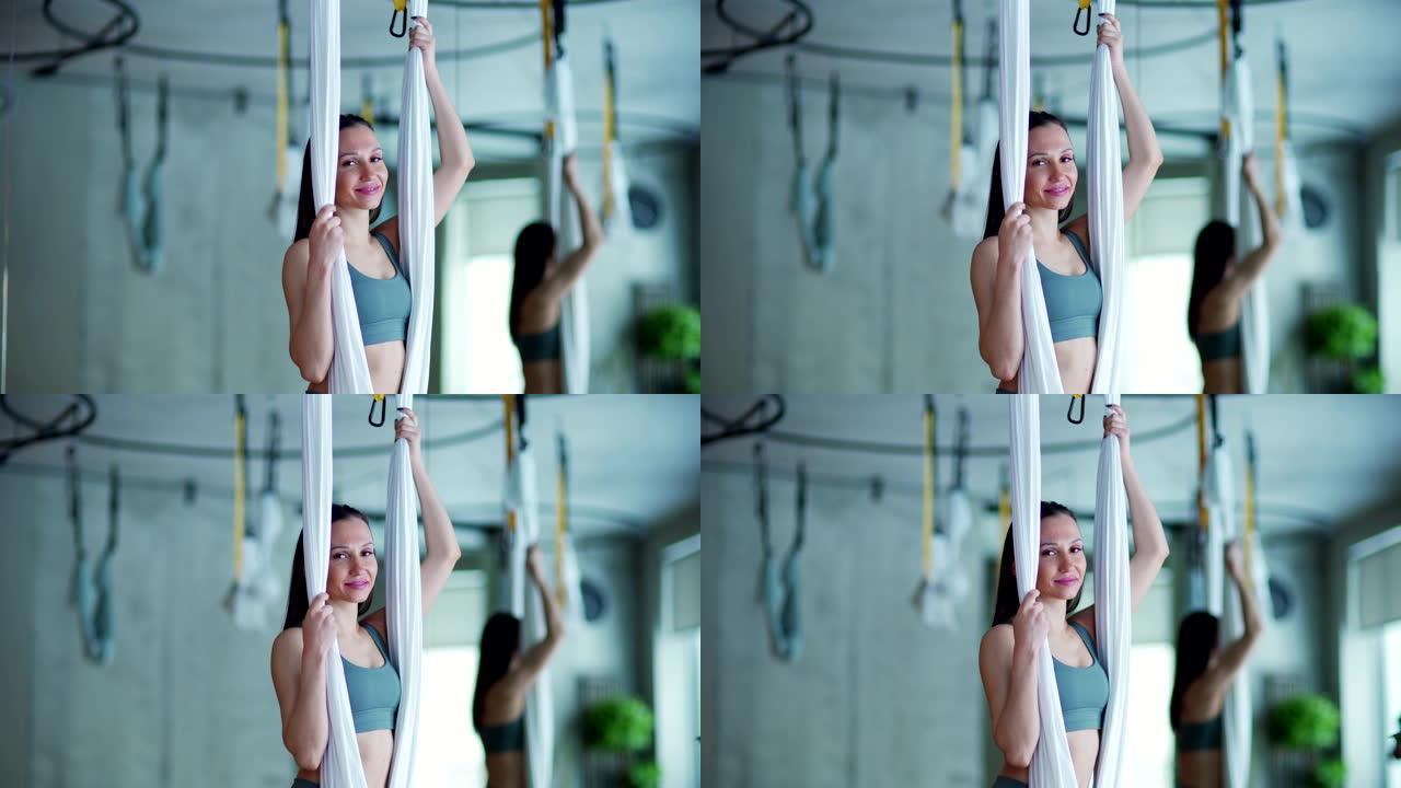 腰部向上的视频肖像，身材健美的年轻女子站在空中瑜伽馆的吊床上看着相机，微笑着