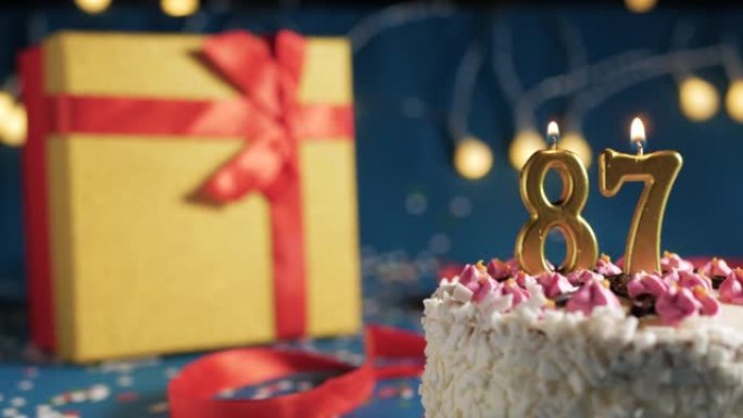白色生日蛋糕87号点火器燃烧的金色蜡烛，蓝色背景带灯和用红丝带捆绑的礼物黄色盒子。特写