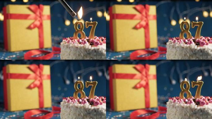 白色生日蛋糕87号点火器燃烧的金色蜡烛，蓝色背景带灯和用红丝带捆绑的礼物黄色盒子。特写