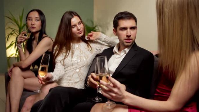 英俊的女性男性坐在沙发上，在派对上与三个漂亮的年轻女性在起泡酒上交谈和调情