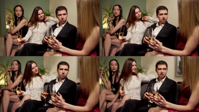 英俊的女性男性坐在沙发上，在派对上与三个漂亮的年轻女性在起泡酒上交谈和调情