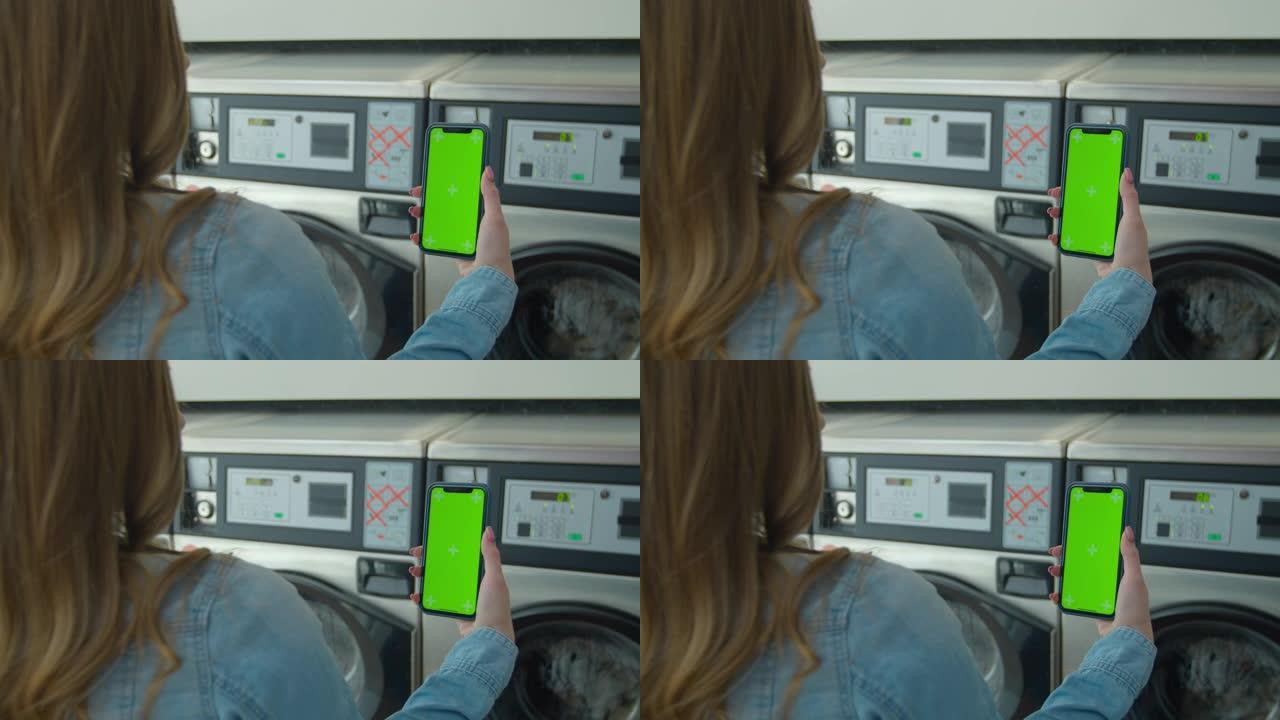 女人在自助洗衣店洗衣服时使用带有绿屏的智能手机。色度键。特写。