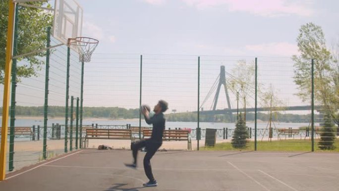 运动型迷人的非洲裔美国男子篮球运动员的肖像把球扔进篮筐，在城市户外的球场上庆祝，背景是桥