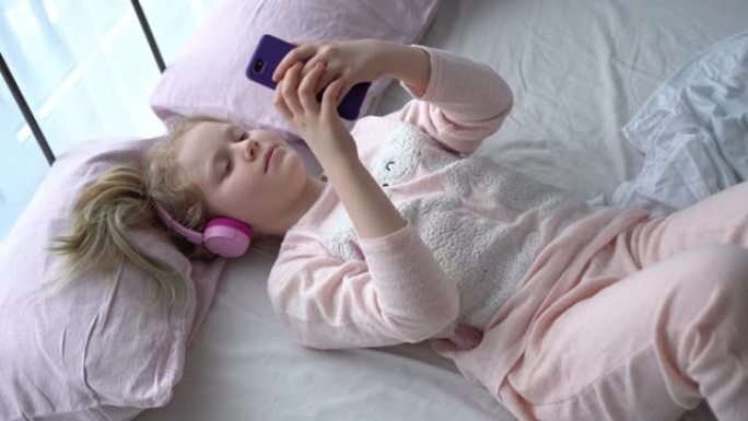 Z世代的现代生活。穿着睡衣和耳机的少女在床上的房间里听智能手机的音乐。