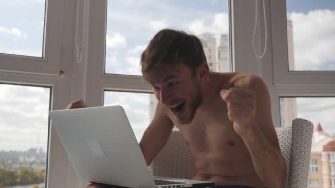 快乐快乐的白人男性玩家穿着内裤在笔记本电脑上玩在线视频游戏并获胜，举起双手欢呼