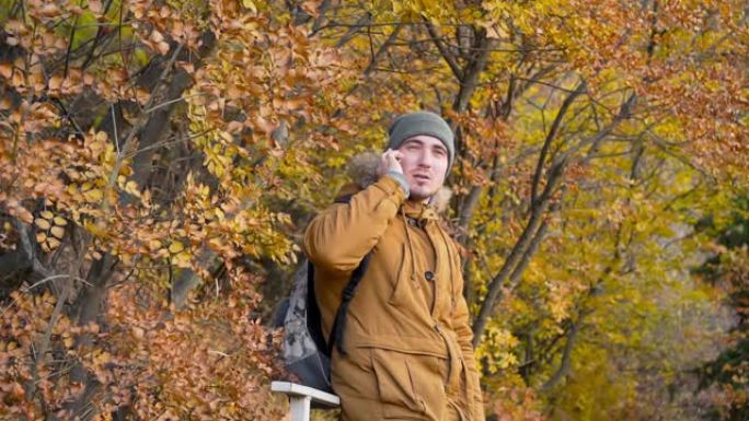 戴着帽子的年轻男性游客，一件带背包的夹克在秋天森林的背景下用智能手机聊天