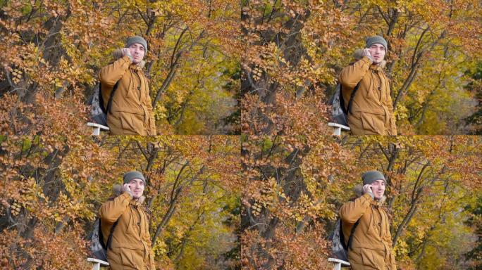 戴着帽子的年轻男性游客，一件带背包的夹克在秋天森林的背景下用智能手机聊天