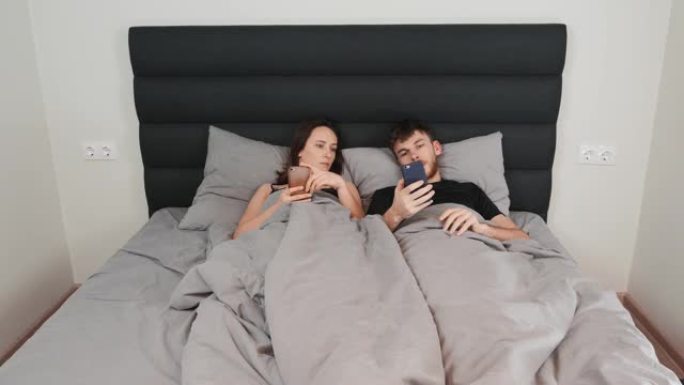 女人和男人躺在床上，手里拿着智能手机。年轻的已婚夫妇在灰色床上观看设备并在网络中发短信。卧室里的男女