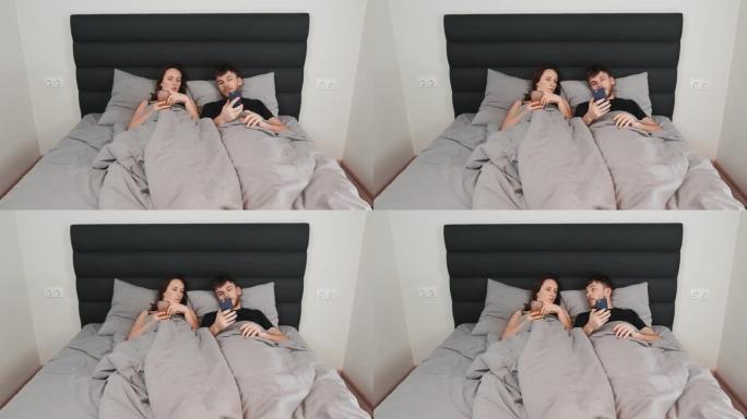 女人和男人躺在床上，手里拿着智能手机。年轻的已婚夫妇在灰色床上观看设备并在网络中发短信。卧室里的男女