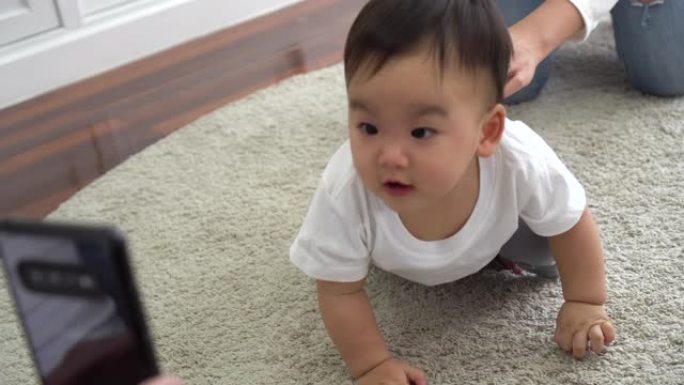 父母使用智能手机拍摄可爱的亚洲婴儿在家中柔软的白色地毯上爬行的视频