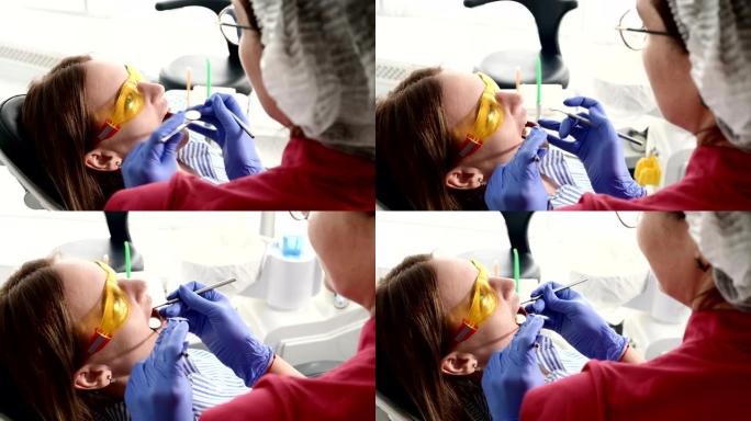 穿着黄色防护眼镜的漂亮金发女孩在踩踏医生检查她张开的嘴。女牙医在牙医仪器的帮助下检查年轻患者的口腔