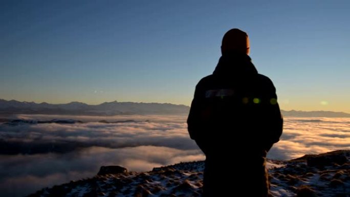 从后面看。一个孤独的站立在高山上的人看着夕阳和日落的地平线，那里充满了云层。旅游旅行与男性孤独的概念