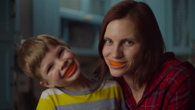 有趣的家庭在家里嘴里玩橘子皮。微笑的女人和学龄前儿童，嘴里有橙色水果。橙色的微笑。