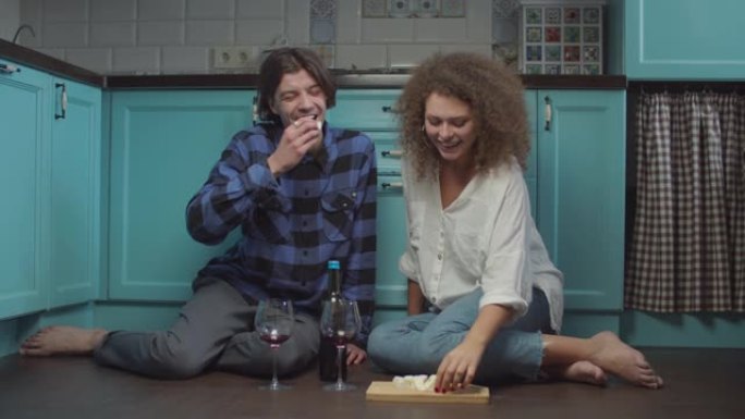 20多岁的年轻夫妇坐在厨房地板上喝酒，吃奶酪。快乐微笑的男人和女人在家喝杯酒庆祝。